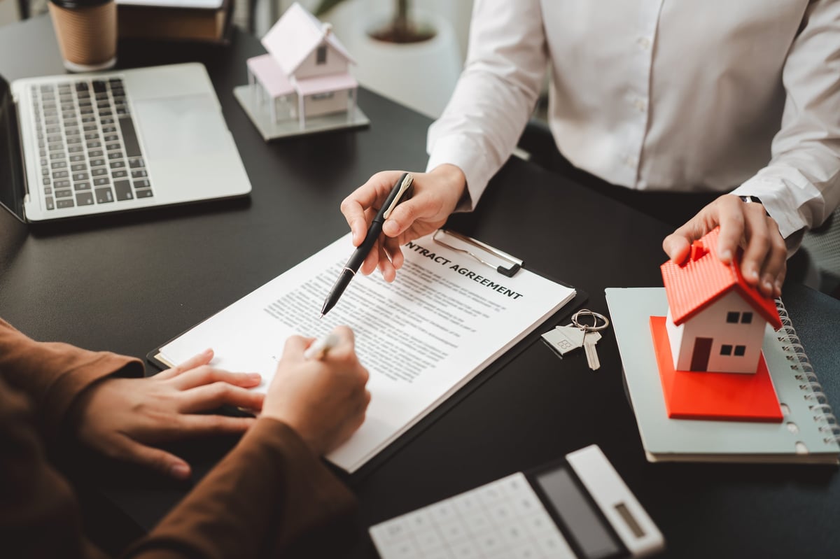 Two people closing a real estate deal at a desk, one signing a contract while the other holds a model house.